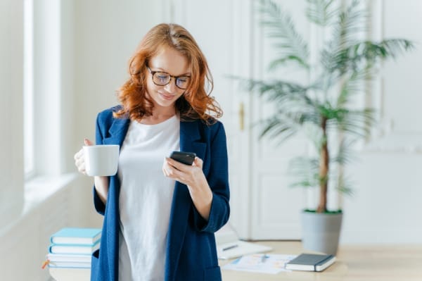 Woman holding a smartphone and a coffee mug, casually checking her phone—representing a user securely connected over Wi-Fi, symbolizing the confidence gained from implementing proper Wi-Fi security measures.